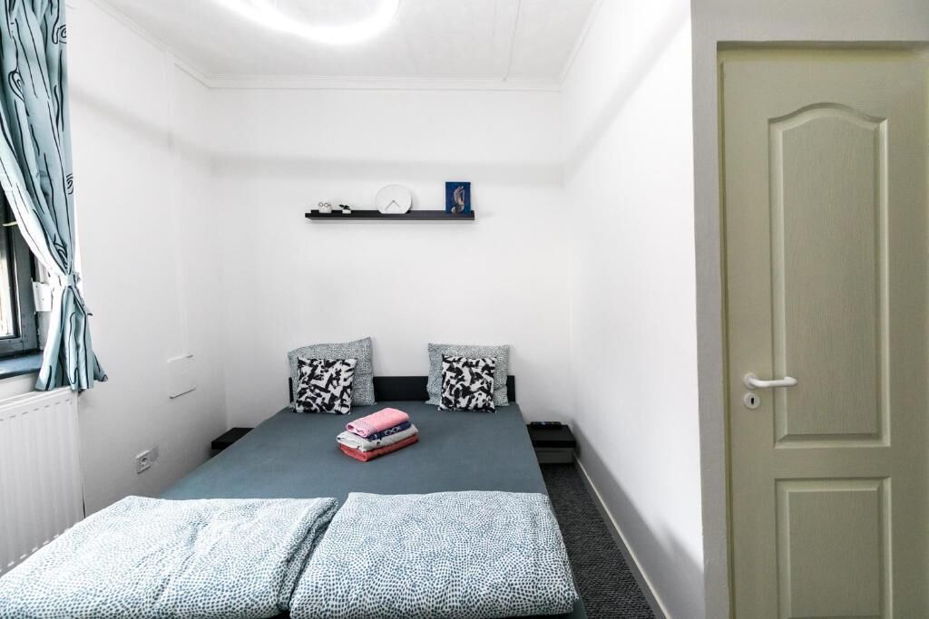 Una habitación pequeña con cama individual, colcha gris, dos almohadas estampadas, un estante con un reloj y un marco de fotos, una puerta y una ventana con cortinas.