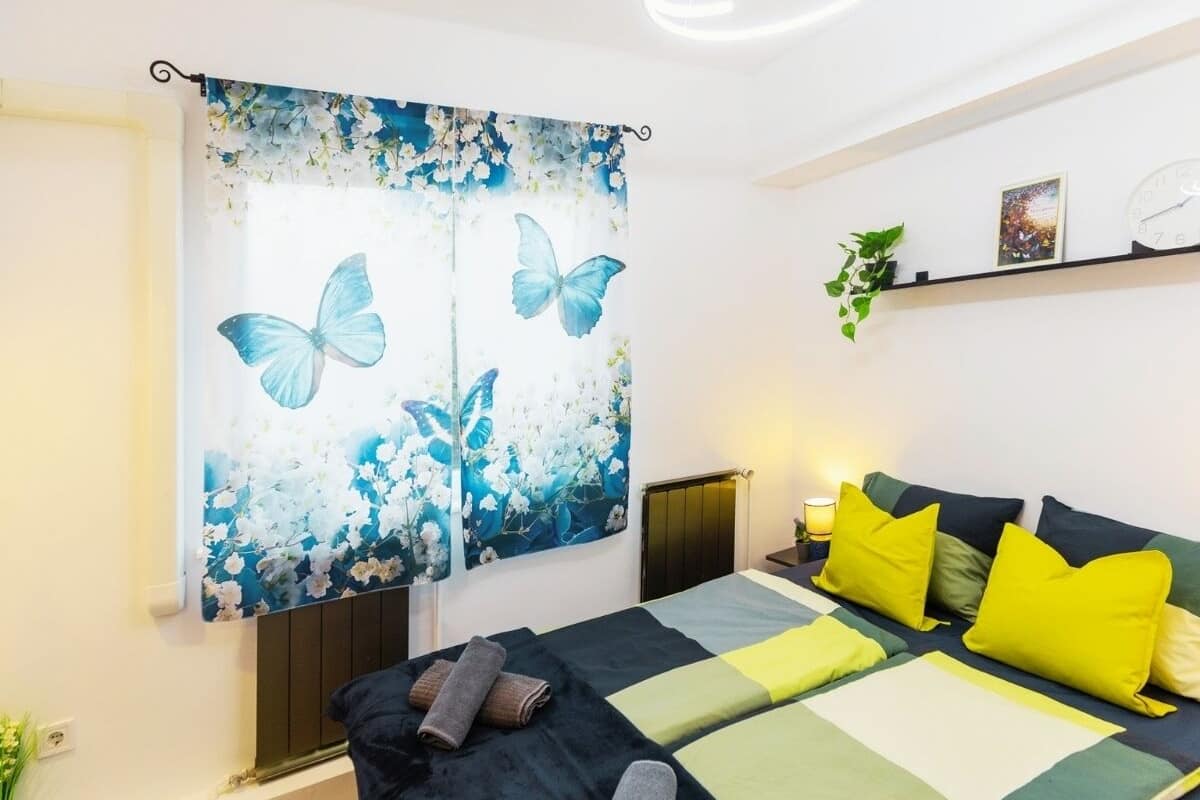 A bedroom with a bed, curtains with a butterfly and flowers, a bedside table with a lamp, and a shelf with a plant.