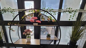 Estante metálico circular con plantas y flores en macetas, frente a una ventana con vista a la ciudad.