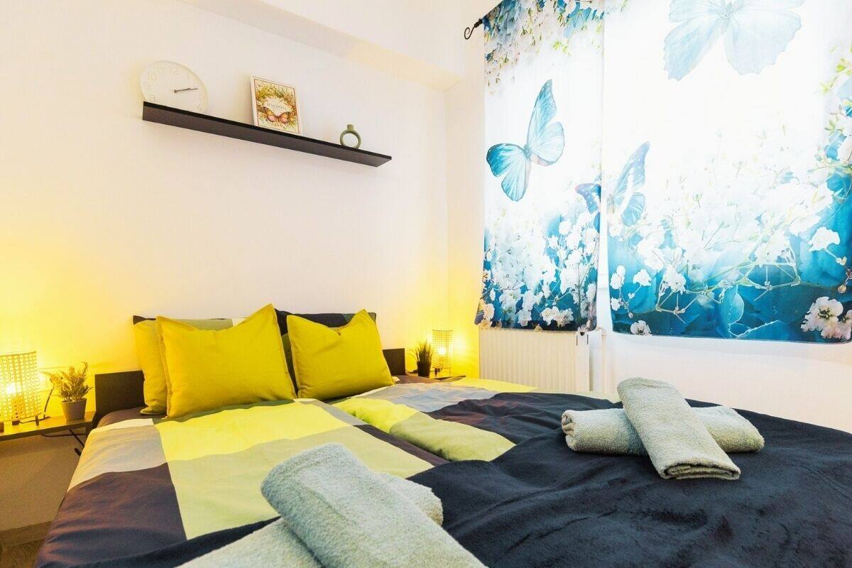 A bed with a black and yellow bedspread, two yellow pillows, and two rolled towels on the bed.