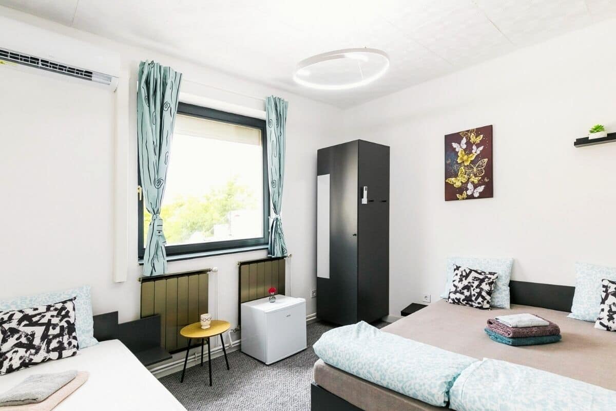 A modern hotel room with a bed, a desk, a chair, a radiator, a wardrobe, and a window with curtains.