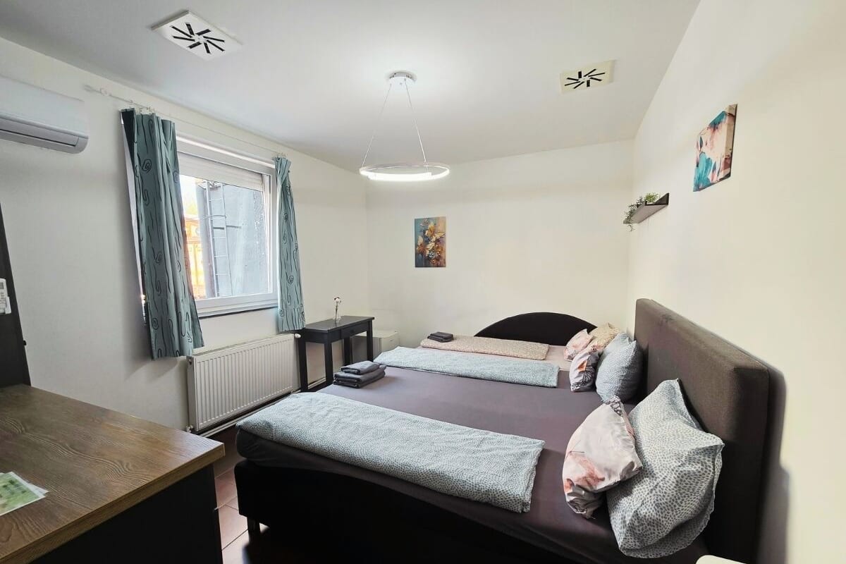 A bedroom with two beds, a desk, a radiator, and a window with curtains.