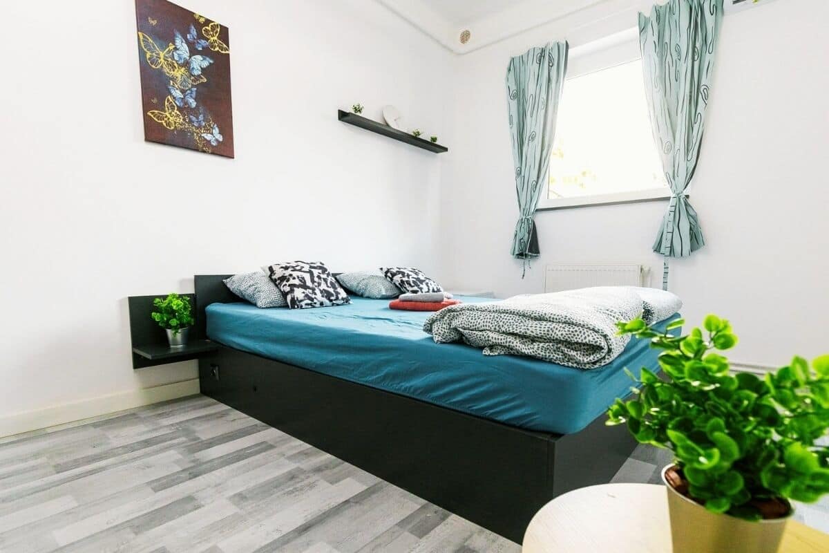 A modern bedroom with a bed, a small table, a potted plant, and a window with curtains.