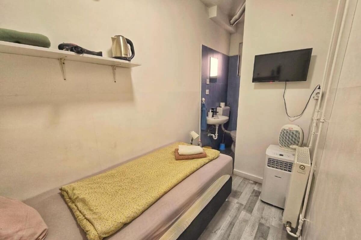 A small, single-bed room with a bed, a shelf with toiletries, a TV, a heater, and a mirror.