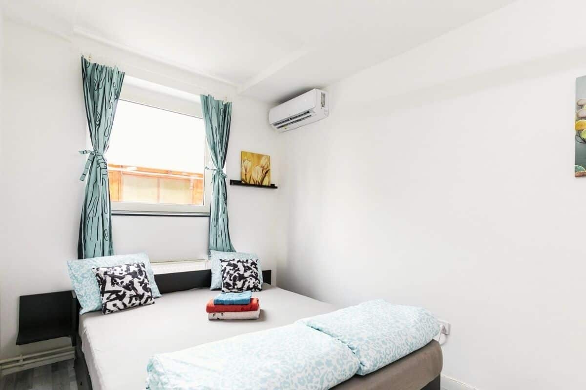 A bedroom with a bed, curtains, an air conditioner, a window, and a painting on the wall.