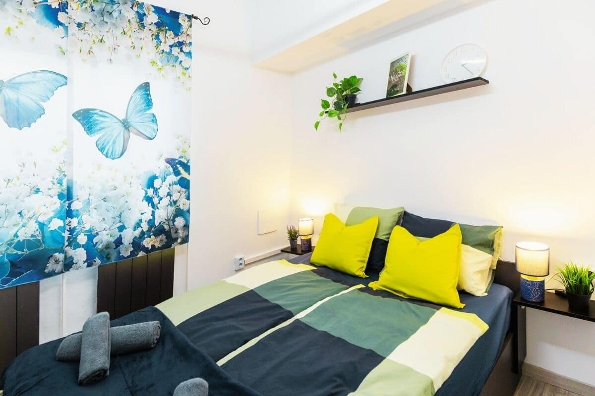 A bedroom with a bed, pillows, a nightstand, a wall clock, a shelf with plants, and a floral tapestry.