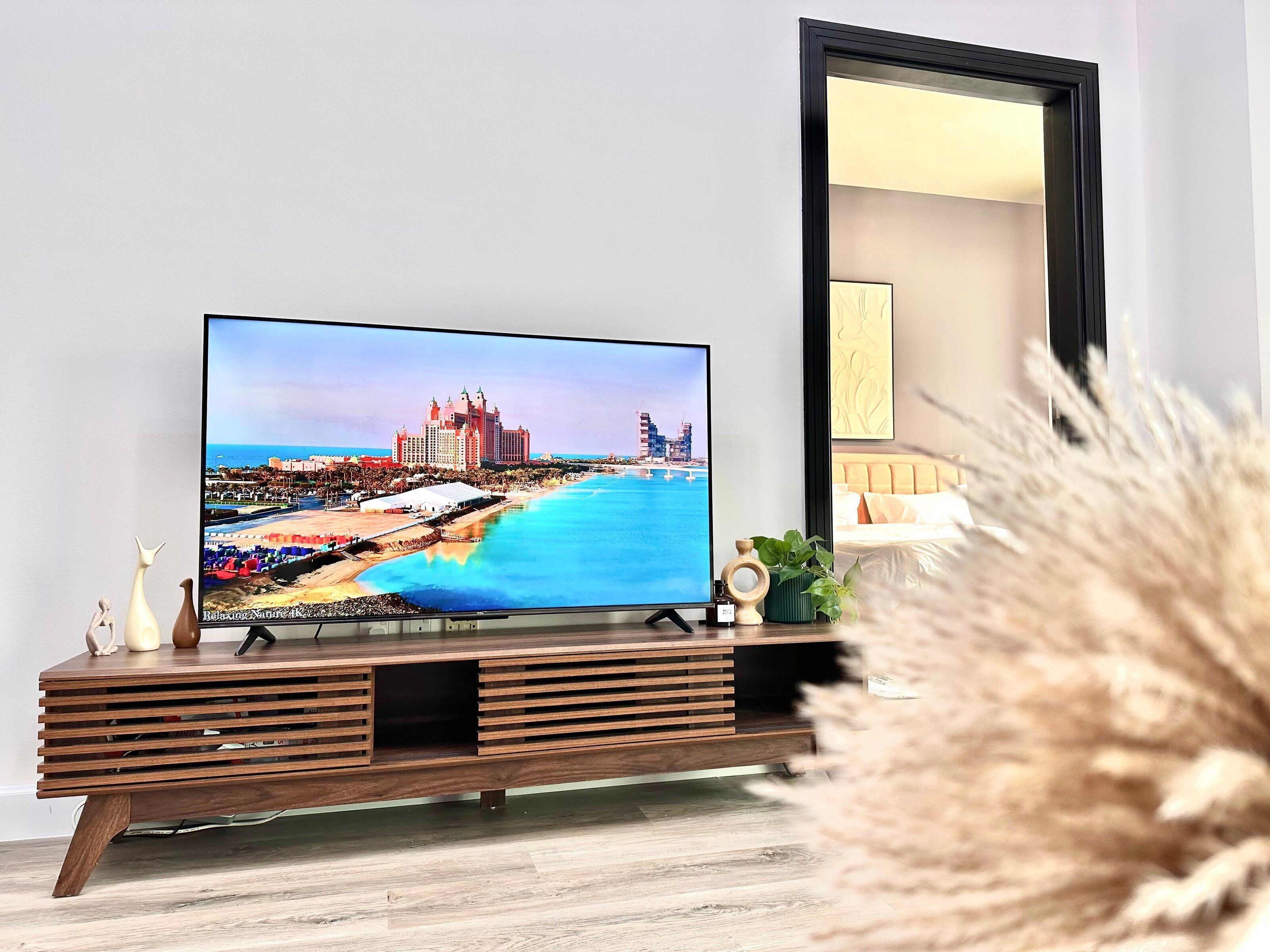 A television on a wooden TV stand with a coastal cityscape on the screen, a mirror, and decorative items.