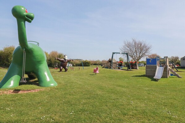 Ein Spielplatz mit einer großen, grünen Dinosaurierskulptur, Schaukeln und einer Rutsche.