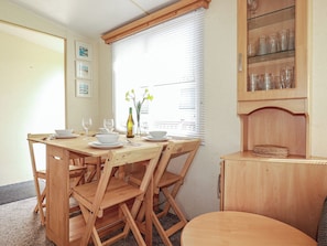 A dining area with a wooden table and chairs, a window with blinds, and a cabinet with glassware. - Beech Lodge (Wadebridge)
