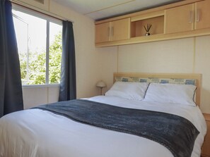 Ein Hotelzimmer mit Bett, einem Fenster mit Vorhängen, einem Nachttisch mit Lampe und hängenden Holzschränken über dem Bett.