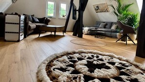 Une pièce de style « loft », avec un plafond haut, des poutres apparentes, un plancher en bois, un tapis rond, un canapé, un fauteuil, une table et une plante.