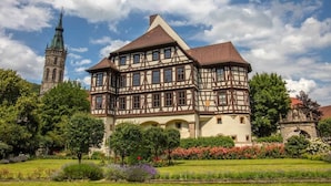 Un grand bâtiment historique, doté d’une tour imposante, entouré de jardins luxuriants et surplombé d’un ciel dégagé.