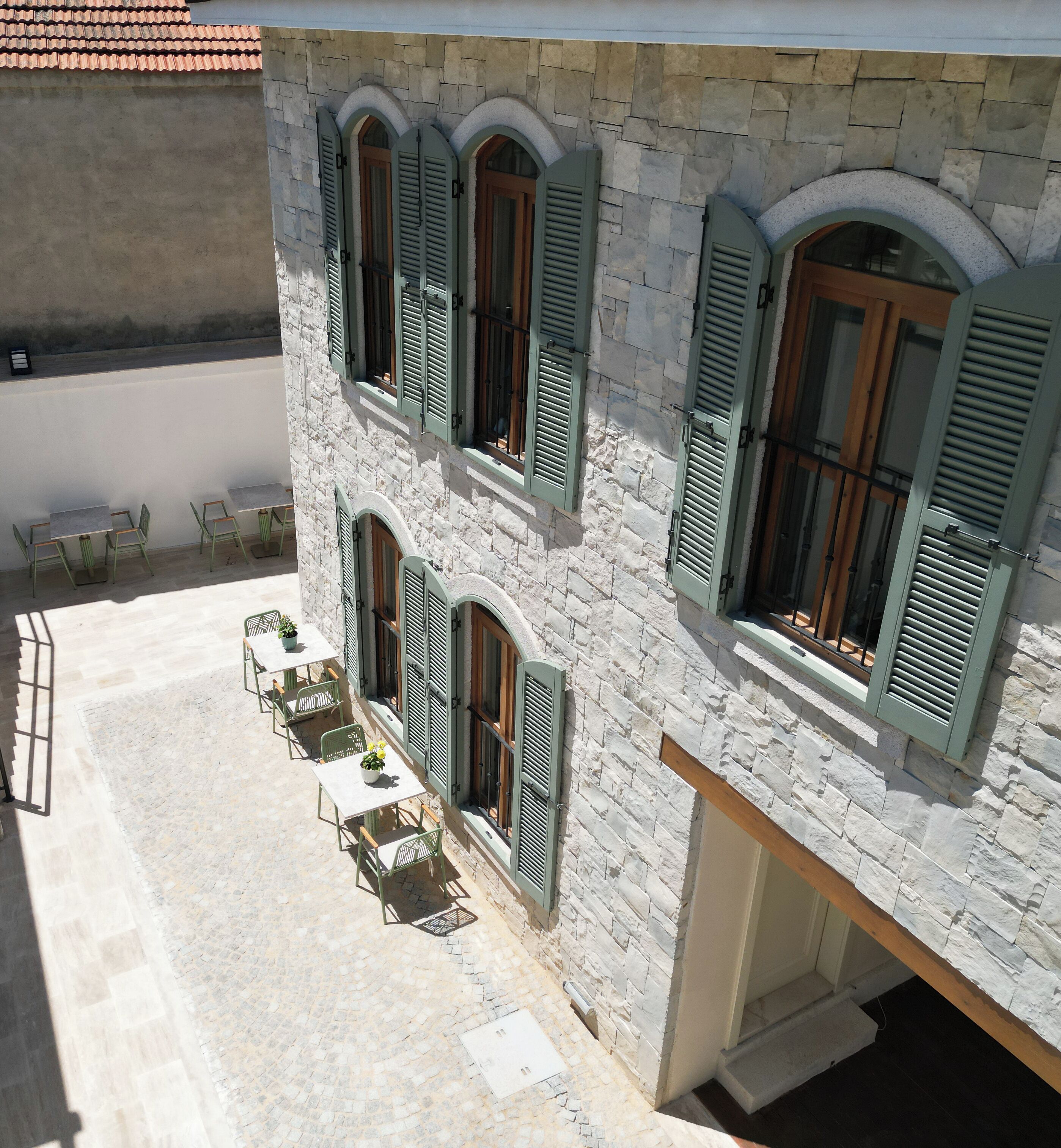 A stone building with arched windows and shutters, outdoor seating with tables and chairs, and a cobblestone patio.