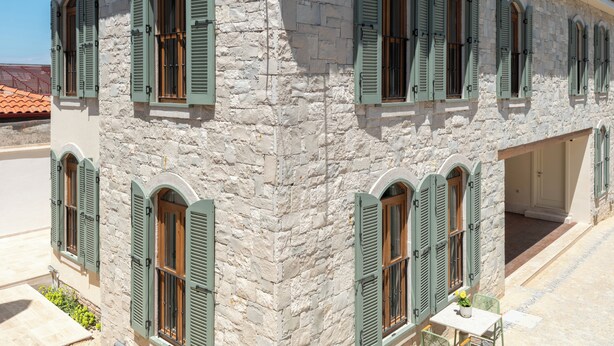 Un bâtiment en pierre doté de nombreuses fenêtres et de volets, d’un toit en tuiles et d’un petit espace extérieur aménagé pour s’asseoir.