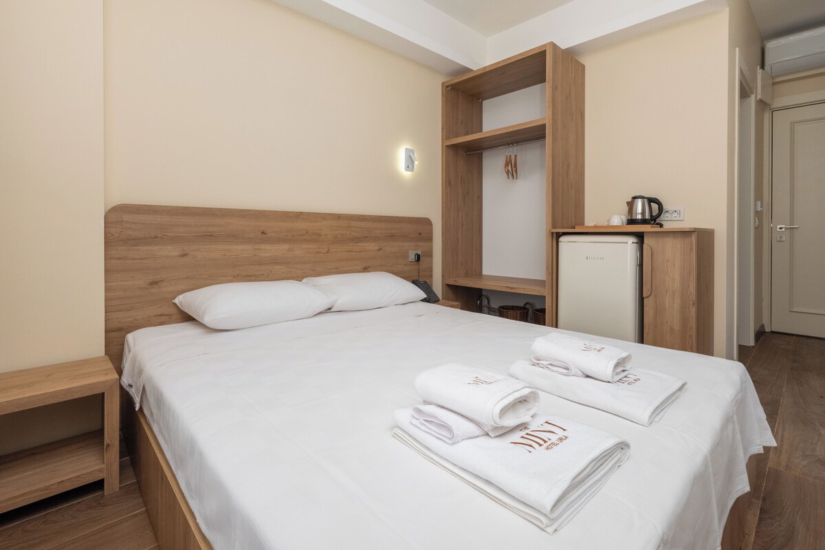 Habitación de hotel con cama de cabecera de madera, armario empotrado, kitchenette pequeña y mesita de noche.