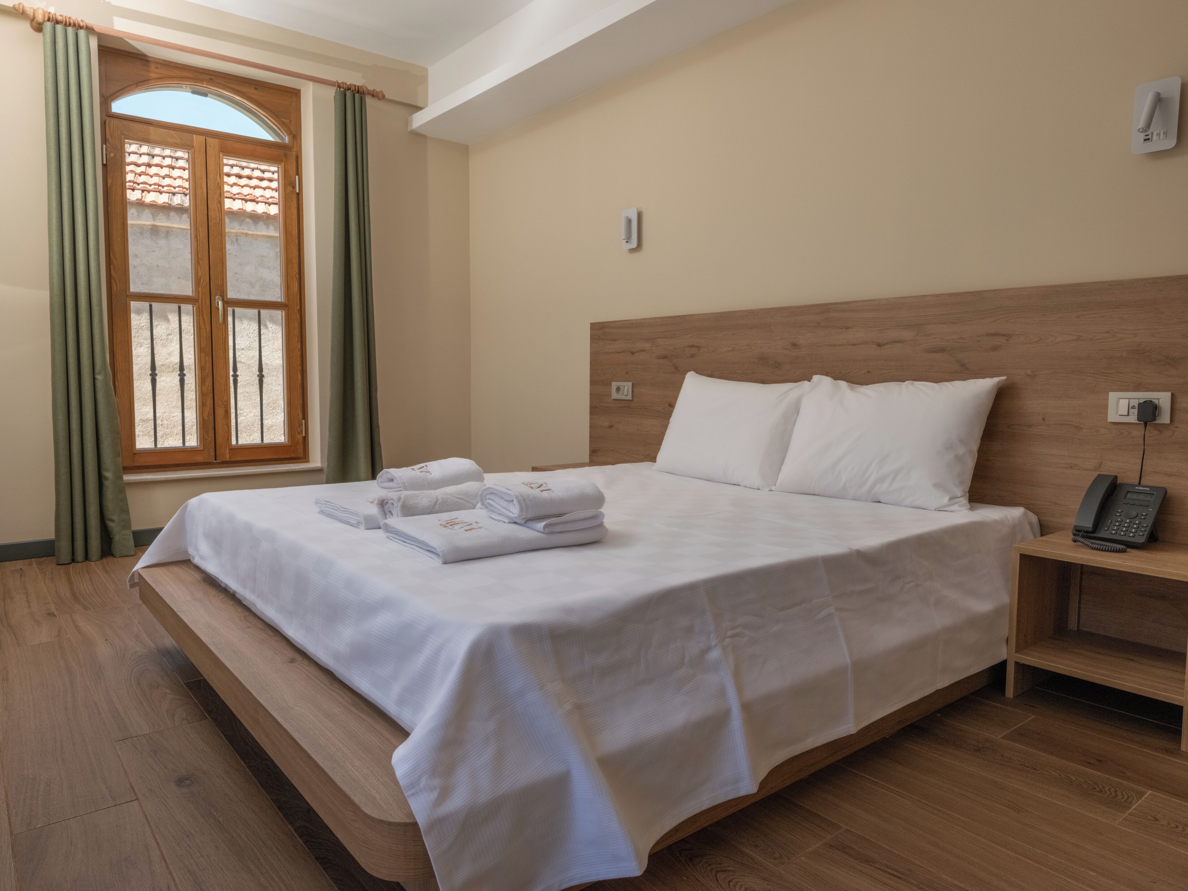 Habitación de hotel con cama, mesita de noche con teléfono, ventana con cortinas y suelo de madera.