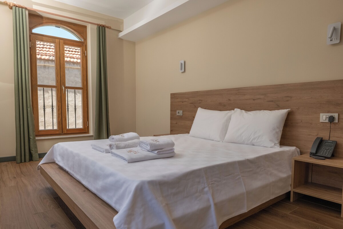 Habitación de hotel con cama, mesita de noche con teléfono, ventana con cortinas y suelo de madera.