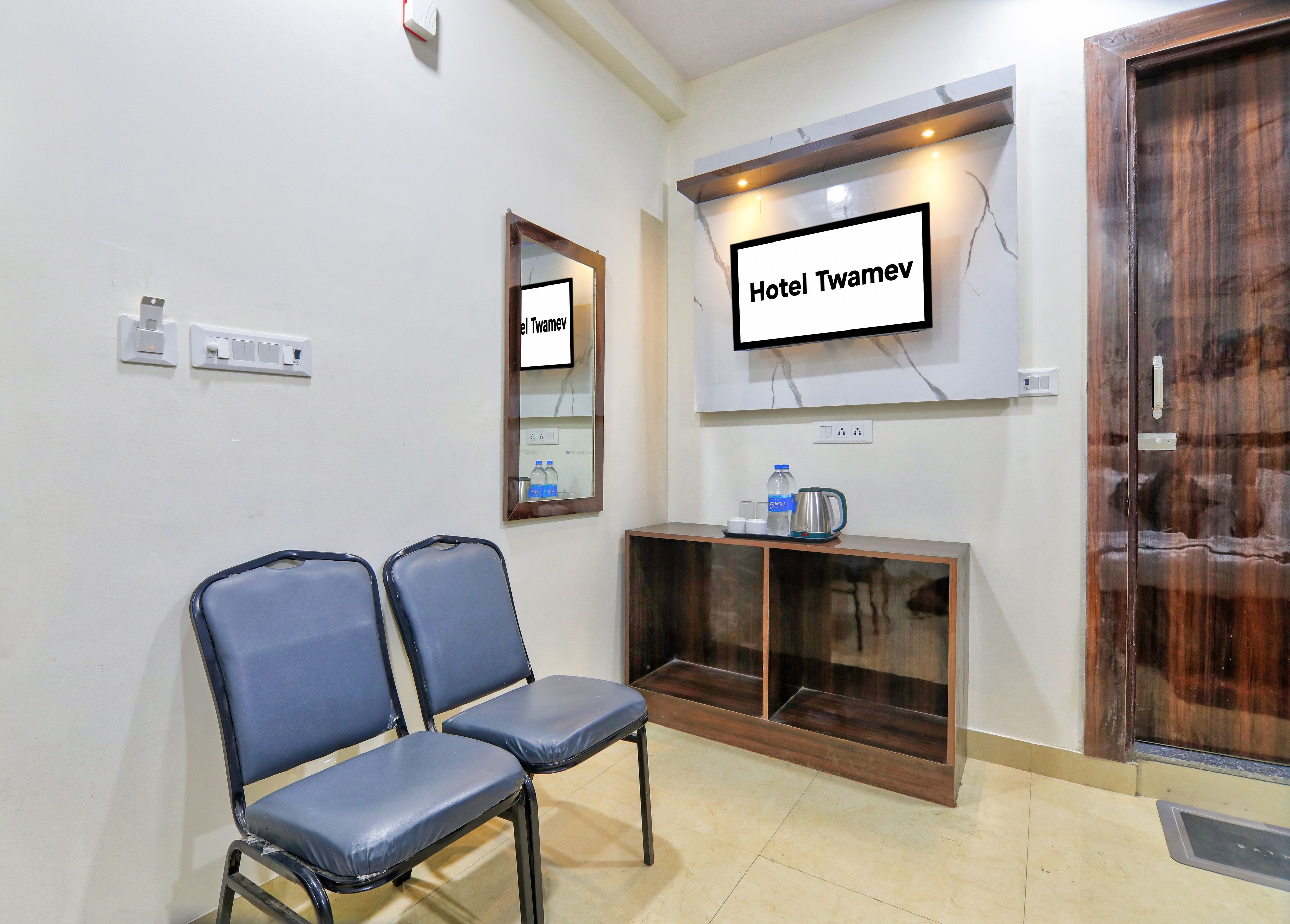 A hotel reception area with a reception desk, two blue chairs, a wall-mounted TV, and a wooden door.