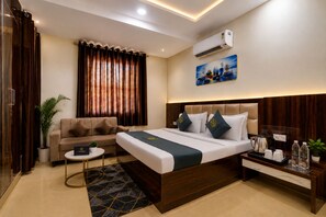 A hotel room with a bed, a sofa, a coffee table, a side table with a kettle, and a potted plant. - Jeet residency by twamev (New Delhi)