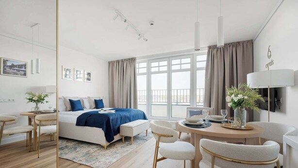 A modern hotel room with a bed, bedside table, lamp, and a view of the city.