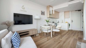 A modern living room with a flat-screen TV, a sofa with patterned pillows, a dining area with a round table and chairs, and a kitchen in the background.
