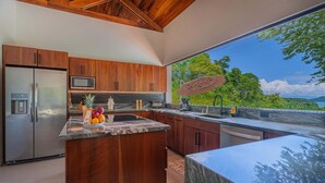 A modern kitchen with wooden cabinets, a stainless steel refrigerator, a microwave, and a large island with fruit. - Modern secluded villa with private pool, jungle setting, and stunning Gulf views near Golfito. (Golfito)
