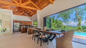 A modern kitchen with wooden cabinets, a dining area with a large table and chairs, and a view of a swimming pool. - Modern secluded villa with private pool, jungle setting, and stunning Gulf views near Golfito. (Golfito)