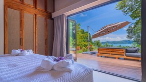 A bedroom with a large bed, a wooden wardrobe, and a view of a balcony with a sofa and umbrella. - Modern secluded villa with private pool, jungle setting, and stunning Gulf views near Golfito. (Golfito)
