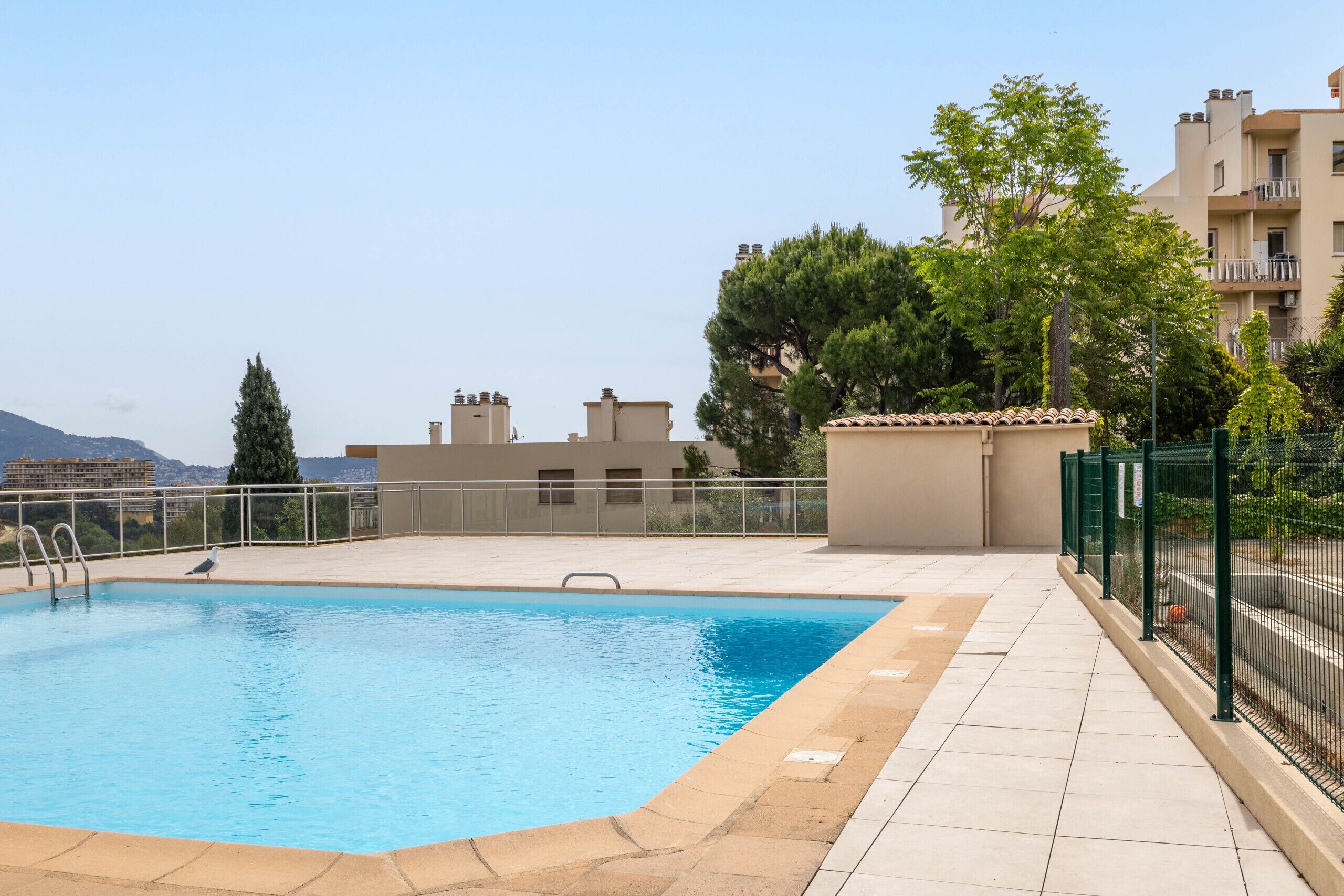 A swimming pool with a tiled deck, surrounded by a fence, and buildings in the background.