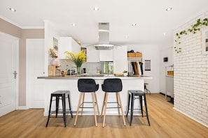 A modern kitchen with white cabinetry, a central island, two black bar stools, and a brick accent wall. - HAPPYFEW SUITE FABRON (Nice)
