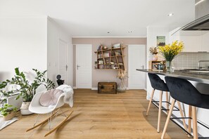 A modern kitchen with a wooden floor, a dining area with black chairs, a white rocking chair, and a bookshelf. - HAPPYFEW SUITE FABRON (Nice)