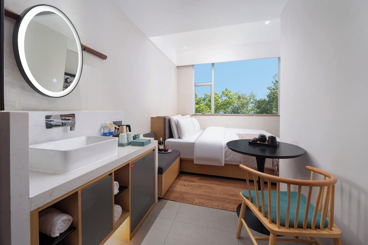A compact room with a bed, a small table, a chair, a round mirror, and a sink.