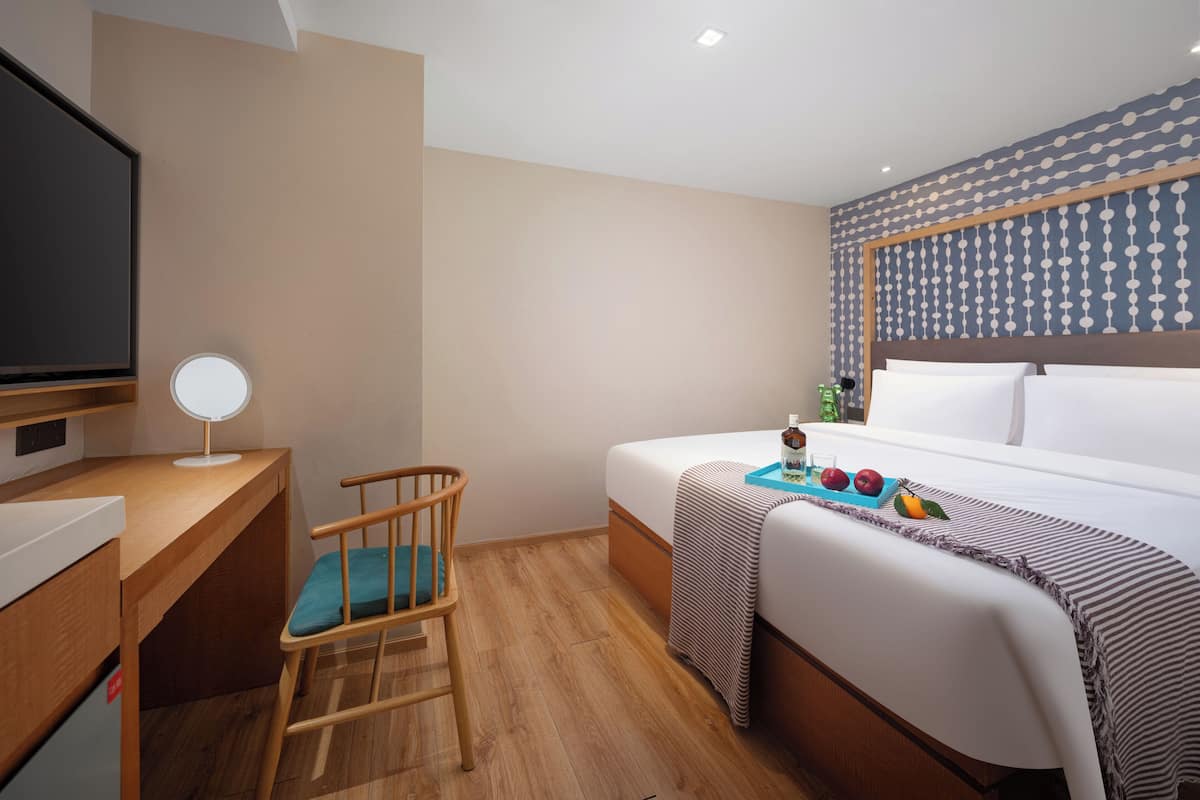 A hotel room with a bed, a desk with a computer, a chair, a TV, and a bedside table.