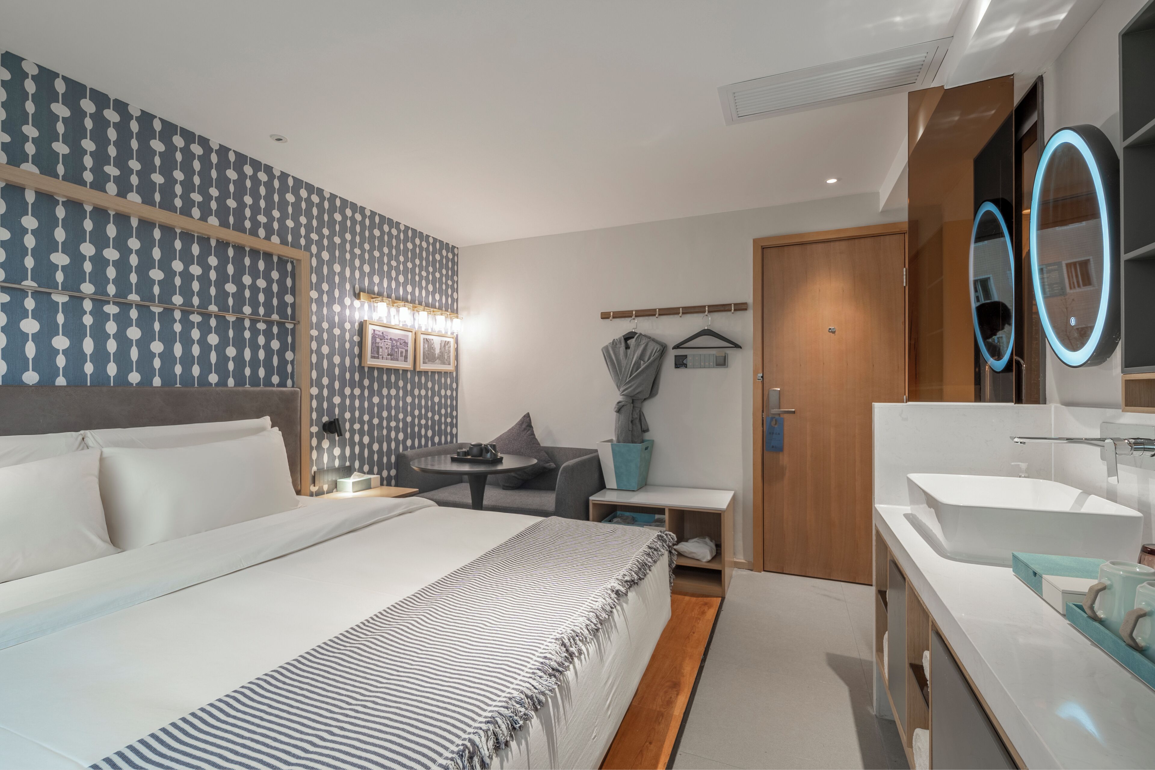 Una habitación de hotel moderna con una cama grande, mesitas de noche, una pequeña mesa redonda, un sofá, un espejo y un baño con lavamanos y espejo.