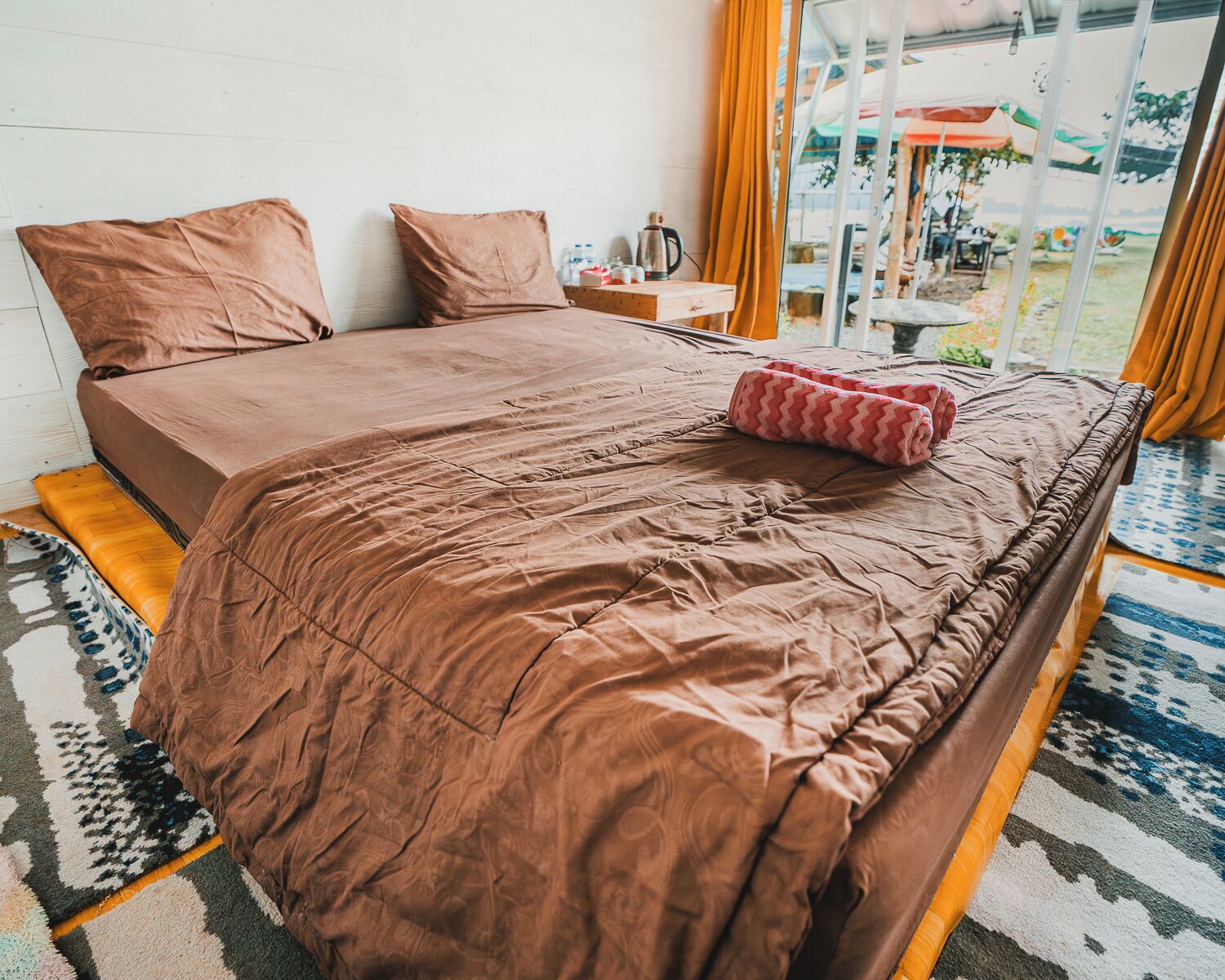 A bed with a brown cover, a pink towel, and a wooden nightstand with a kettle.