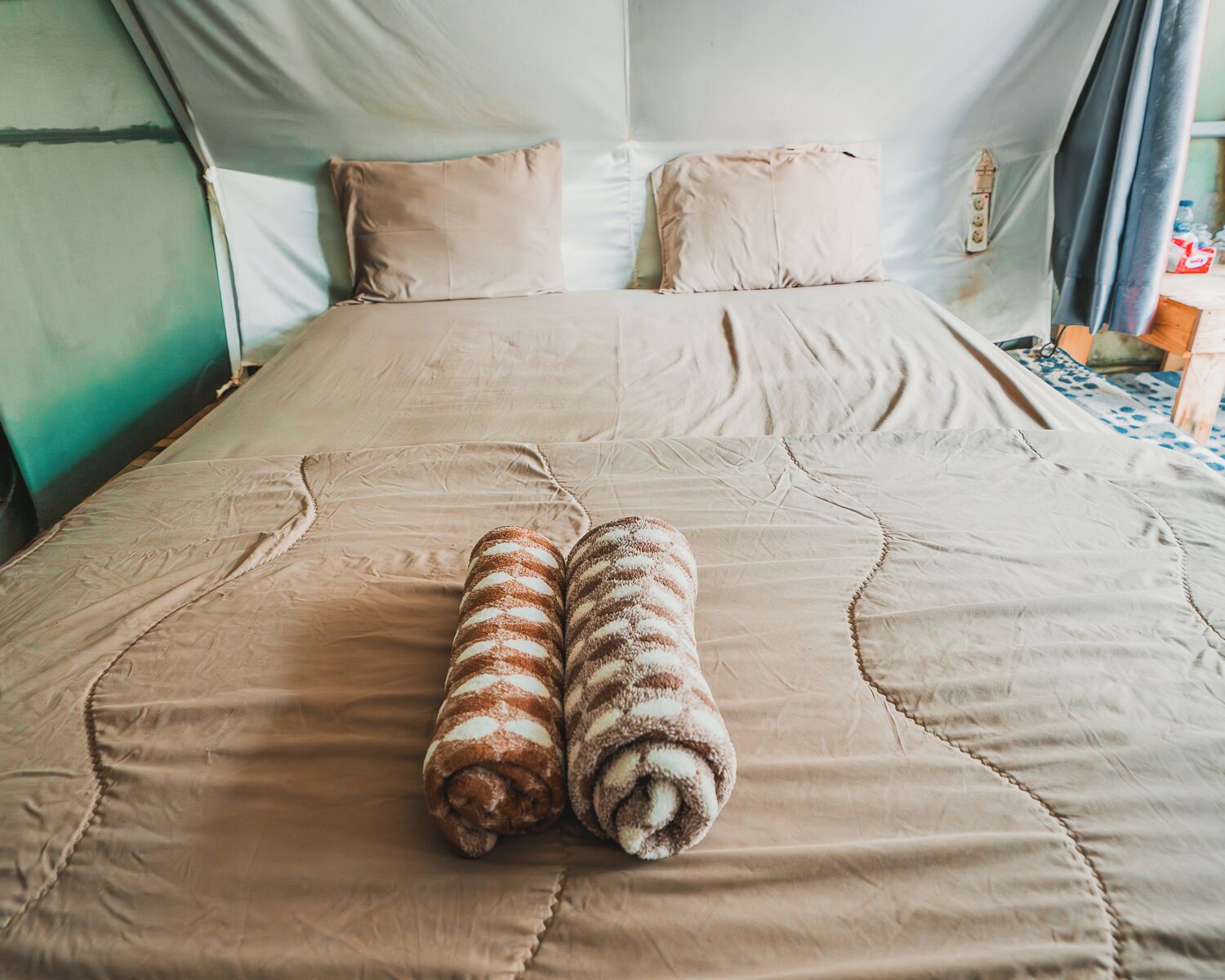 A bed with a beige bedspread, two rolled towels, and two pillows.