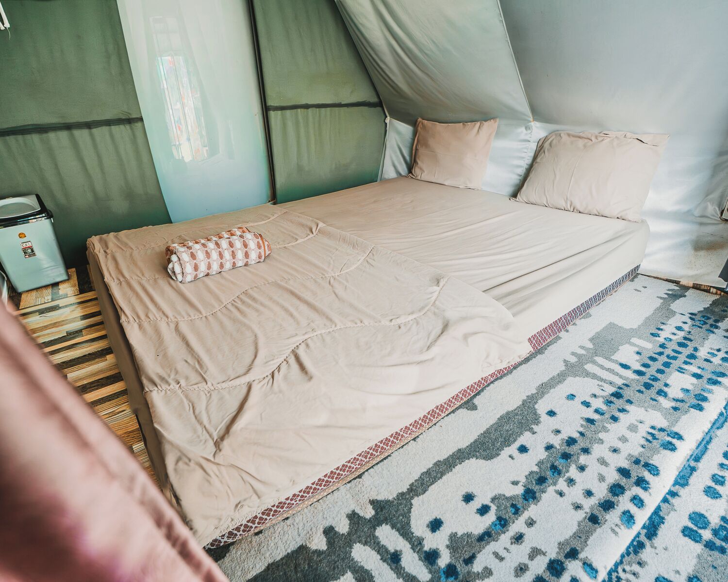 Una cama con una colcha color beige, almohadas, una manta estampada y un tapete con diseño.