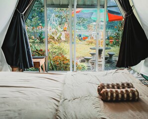 A tented room with a bed, a pillow, and a view of a garden with plants and a fountain. - Catur Glamping Bedugul (Baturiti)
