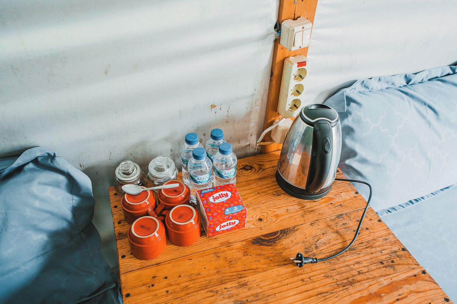 A wooden table with a kettle, water bottles, and orange containers, likely for food storage, against a corrugated metal wall.