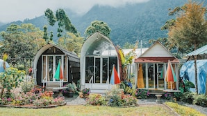 Three small, single-story cabins with large windows, surrounded by a garden with various plants and flowers, set against a mountainous backdrop. - Catur Glamping Bedugul (Baturiti)