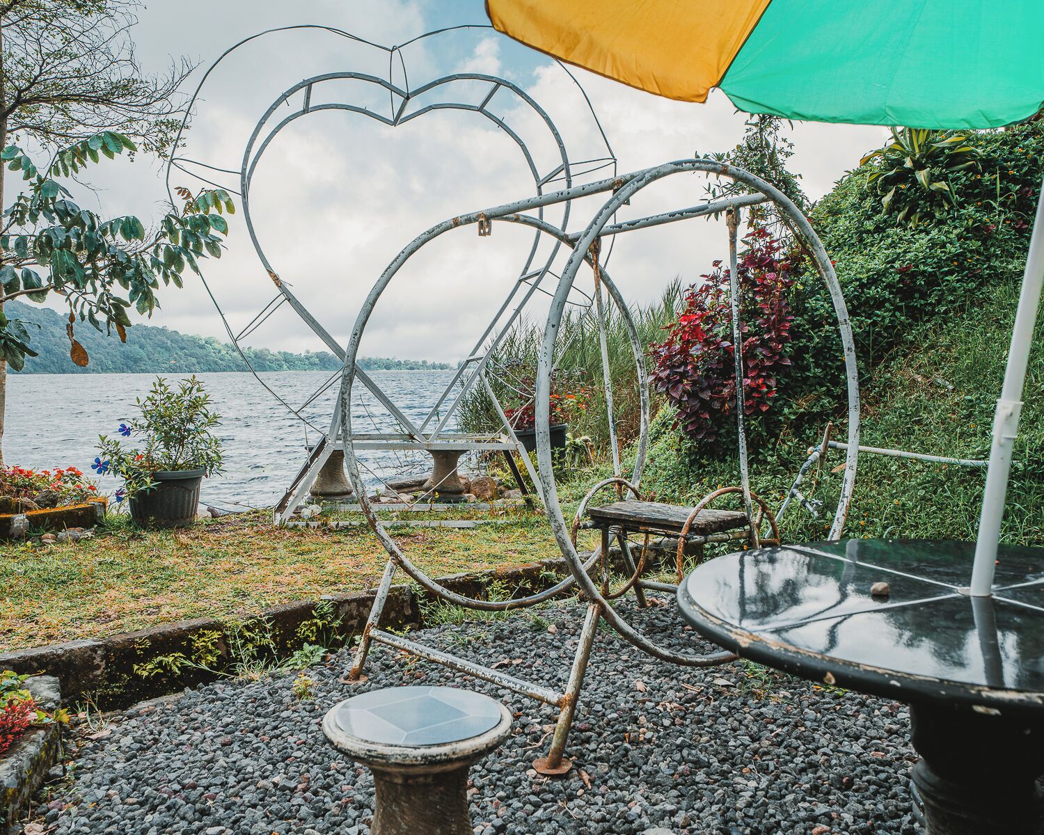 A heart-shaped metal structure with a table and chairs, surrounded by greenery and a body of water.