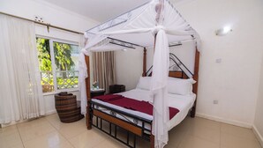 Una cama con dosel, una mesita de noche de madera, un sillón de mimbre y vistas al verde exterior.