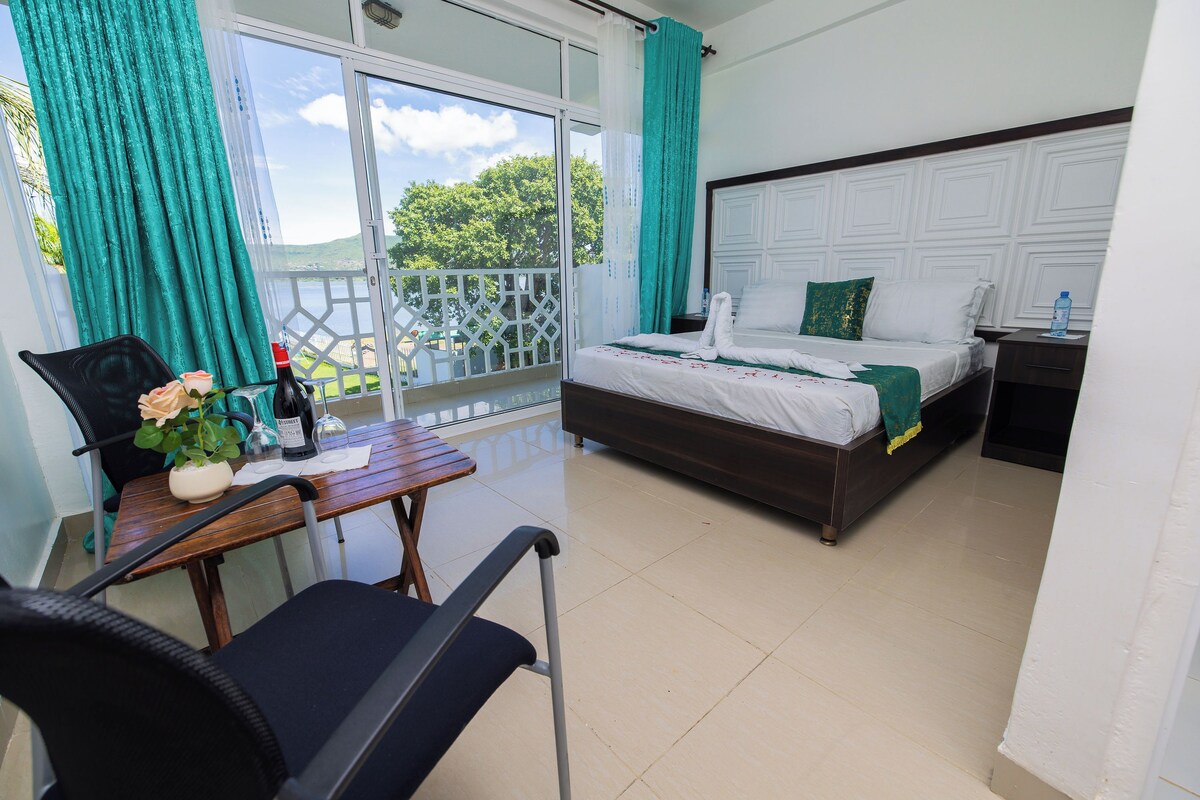 A hotel room with a balcony, a bed with white bedding, a wooden table with a chair, and a potted plant.