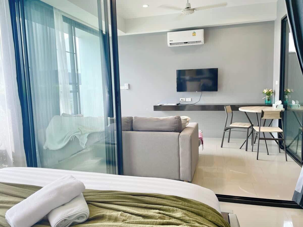 A hotel room with a bed, a sofa, a dining area with a table and chairs, a TV mounted on the wall, and an air conditioning unit.