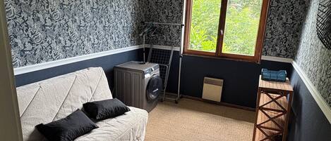 A compact room with a washing machine, a small table, a radiator, and a window with a view of greenery.