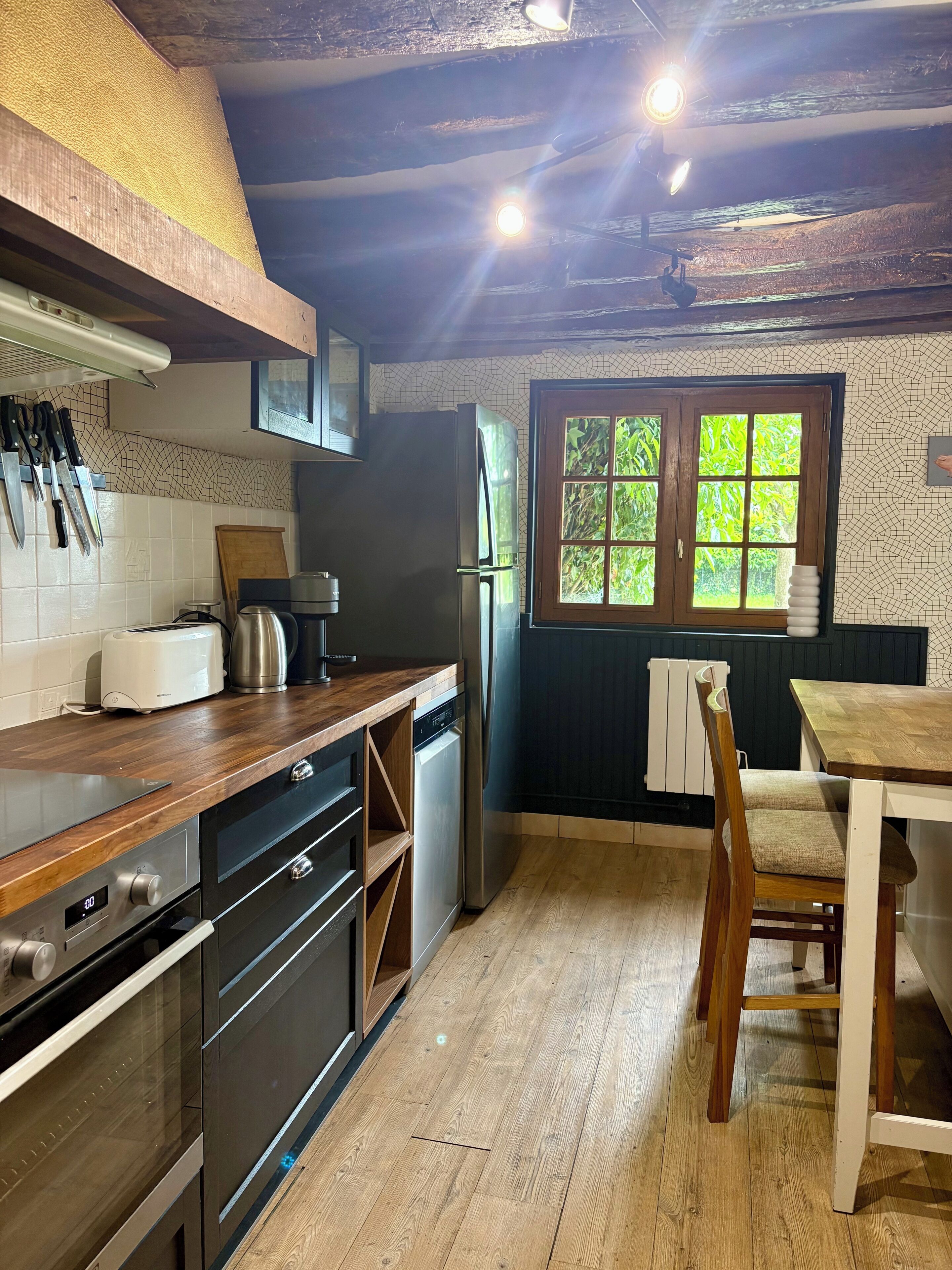 Una cocina con armarios de madera, un refrigerador de acero inoxidable, una mesa de comedor de madera y una ventana con vista a vegetación.
