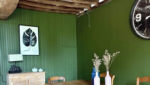 Een rustieke eetruimte met een houten tafel, stoelen, een groene wand, een klok en een kunstwerk met bladeren in een lijst.