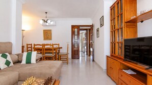 Uma sala de estar com sofá, mesa de centro com xadrez, televisão, armários de madeira, área de refeições e corredor.
