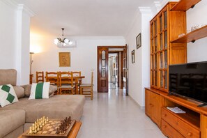 A living room with a sofa, a coffee table with chess, a TV, wooden cabinets, a dining area, and a hallway. - 284 La Santa by Mallorca Charme (Ca´n Picafort)
