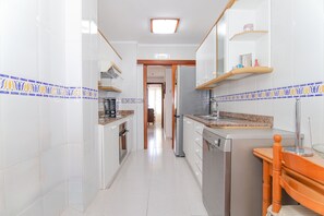 A narrow kitchen with white cabinets, a granite countertop, and a wooden chair. - 284 La Santa by Mallorca Charme (Ca´n Picafort)